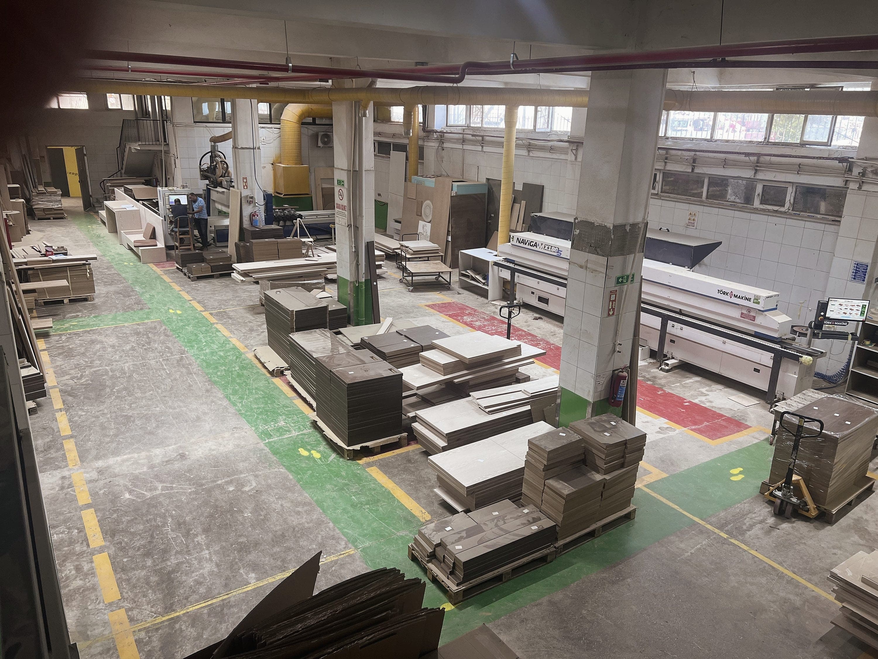 Die Halle: Die Produktionshalle von oben: Plattenstapel auf Paletten, die langen Maschinen an der hinteren Wand.