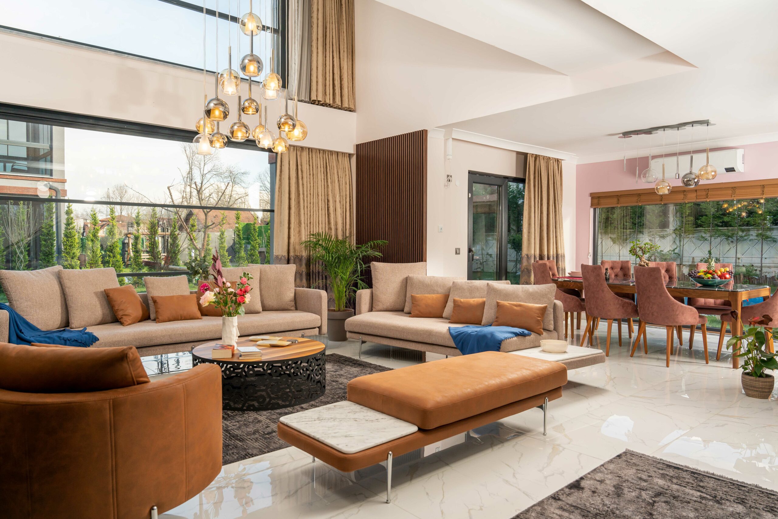 Sapanca villa great room furnished by Naggarco: double-height living and dining under a cascading glass chandelier, cognac leather daybed with marble ends, sofas and dining seating facing the garden glazing.